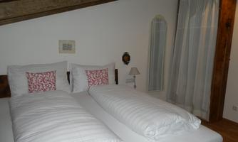 A cozy bedroom with two single beds and elegant cushions. The walls are light, and a curtain lets soft light in.