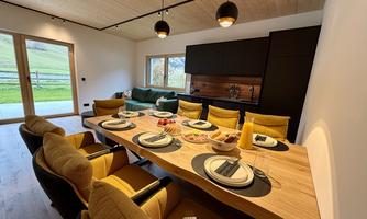 A modern dining room with a large table and comfortable chairs. In the background, there is a green couch and windows overlooking the outdoors.