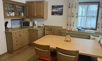 A cozy kitchen with wooden cabinets and a large dining table.  
Curtains with a floral pattern and a view of the countryside complement the ambiance.