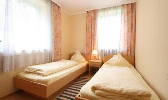 A simple room with two single beds and a bedside table. Daylight streams through the windows with curtains in soft colors.