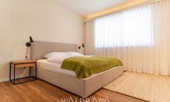 A modern bedroom with a cozy bed and a green blanket. Large windows let in plenty of daylight and the floor is made of wood.