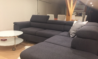 A modern living room with an elegant gray sofa and a round table. The decor is bright and minimalist, featuring wooden sculptures and soft lighting.
