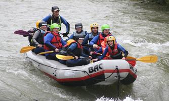 A group of nine people paddles in an inflatable boat on a river. Everyone is wearing life vests and helmets, and the scene conveys a sense of adventure and teamwork.