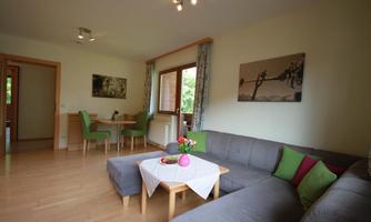A cozy living room with a gray sofa and a small table. The room has large windows and a dining area with green chairs.
