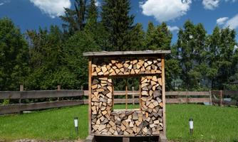 A wooden wood storage made of wood with stacked firewood. In the background, trees and a blue sky are visible.