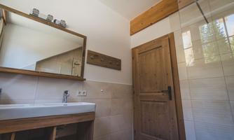 A modern bathroom with a wooden washbasin and a large mirror. The walls are tiled in light colors, and a wooden door leads outside.