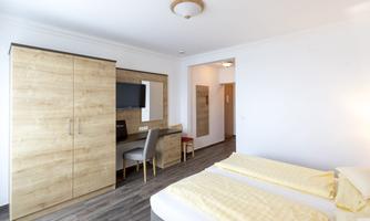 A modern bedroom with two beds, a desk, and a television. The decor is bright and minimalist, featuring wood and neutral tones.