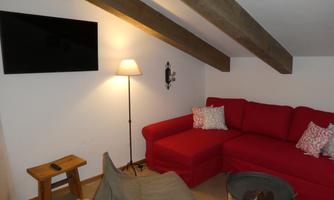 A cozy living room with a red sofa and a standing lamp. A television is mounted on the wall, and there is a small wooden table.