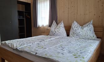 A cozy bedroom with a large bed and bright bed linens. The walls are made of wood, and there is a wardrobe as well as curtains.