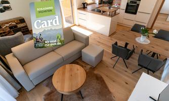 A modern living room with a bright sofa and a round coffee table. In the background, there is an open kitchenette and plenty of daylight.