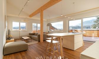 A modern and bright living room with large windows and an open kitchen area. The decor is simple and elegant, featuring wooden elements and a cozy seating area.