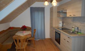A cozy kitchen corner with a wooden cabinet, a small seating group, and a table. The space is bright and friendly, with a window and modern kitchen utensils.