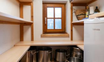 A well-organized pantry with shelves and a window. On the floor are large cooking pots and a fridge-freezer combination.