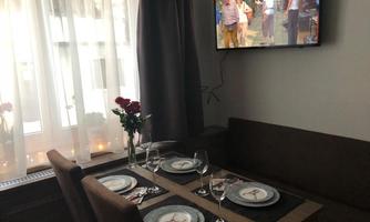 A cozy dining area with a beautifully set table. In the background, a television is playing, and light and plants are streaming in through the window.