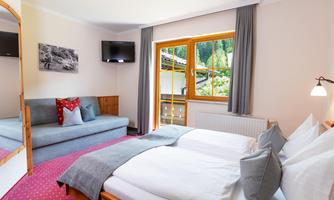 A cozy bedroom with two beds, a television, and a sofa. Large windows offer a view of a green environment.