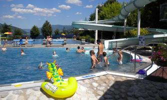A pool with many children swimming and playing. In the background, there is a slide and a beautiful landscape with trees.