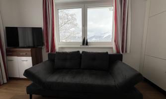 A modern living room with a gray sofa and a television. Through the window, a view of mountains and a cloudy sky is visible.