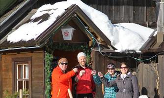 A group of four people stands in front of a house in the snow, decorated with a sign "Roßstall" and a sign featuring the "Siegel" logo. In the foreground, a sled and an orange cat can be seen.