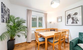 A bright dining room with a wooden table and chairs. A plant stands in the corner, and large windows provide plenty of daylight.