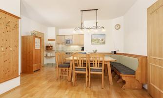 A cozy dining area with a large wooden table and chairs. The kitchen is modern and bright, with wooden shelves on the wall.