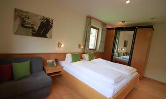 A cozy bedroom with a double bed and a dark blue couch. The room features a large window and a wardrobe.