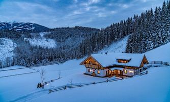 A cozy wooden house in the snow with warm light, surrounded by snow-covered fir trees. The winter landscape stretches out to the mountains in the background.