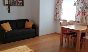 A bright living room with a dark sofa and wooden floor. A dining table with chairs and colorful curtains make the room cozy.