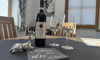 A table setting with a bottle of red wine, two wine glasses, and a silver deer figurine. A placemat with the inscription "Nice to have you here!" is also underneath.