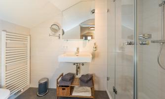 A modern bathroom with a large shower, a sink, and a mirror. Towels are stored in a basket and a towel warmer is mounted on the wall.