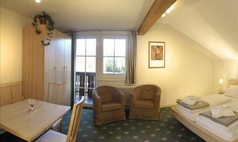 A cozy room with a double bed, two armchairs, and a dining area. Large windows provide light and a view of the outdoors.