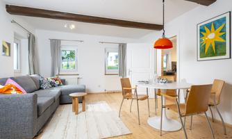 A bright living room with a gray sofa and a round table. The room has large windows and a cozy atmosphere.