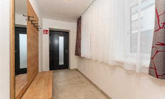 A bright hallway with a wooden cupboard and mirror. The windows have light curtains and there is a black door in the background.