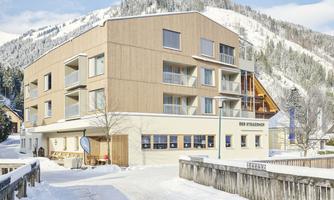A modern building in the snow with large windows and an inviting entrance. In the background, snow-covered mountains rise up.