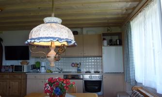 A cozy kitchen with wooden cabinets and a large dining table. Above the table hangs a decorative lamp, and there are fresh flowers in a vase.