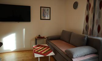A cozy living room with a gray couch and a small table. A television is mounted on the wall, and there is a round wall clock.