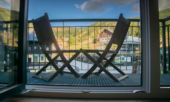 A balcony with two wooden loungers and a view of green mountains. The sky is clear and there are some clouds.