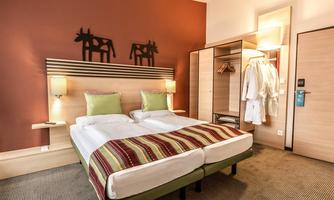 A cozy hotel room with a double bed and colorful cushions. In the background, there is a wardrobe and bathrobes visible.