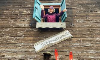 A cozy wooden house with an open window. A person is waving from inside and skis are in front of it.