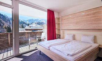 A modern bedroom with a double bed and a beautiful view of the mountains. Large windows and a balcony provide plenty of natural light.