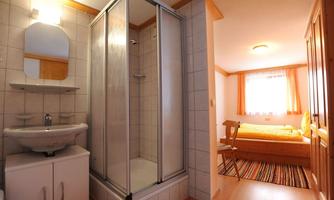 A modern bathroom with a shower cubicle and a sink. In the background, a cozy bedroom with a bed and a window is visible.