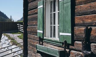 A rustic wooden house with one window and green shutters. The surroundings are rural with a stone wall and trees in the background.