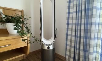 A modern humidifier stands in a bright room. There are wooden furniture and a colored curtain.