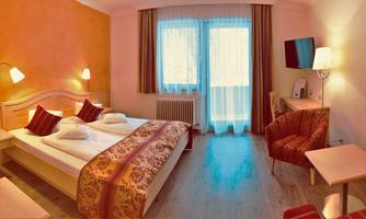 A cozy hotel room with a double bed and a comfortable armchair. The room has large windows with plenty of daylight and a warm color scheme.
