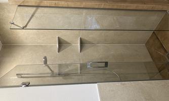 A modern shower with glass walls and an elegant showerhead. The walls are in neutral colors and there are hanging shelves.