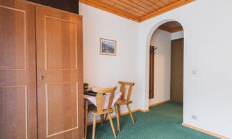 A cozy room with a wooden wardrobe and a table with chairs. The walls are light-colored and an archway leads to another area.