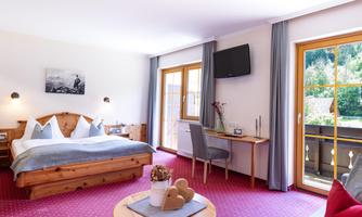 A modern hotel room with wooden details, a double bed, and a desk. Large windows offer a view of the balcony and the natural surroundings.