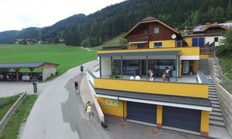 A modern, yellow building in a rural setting. People are walking by on the street, surrounded by green meadows and mountains.
