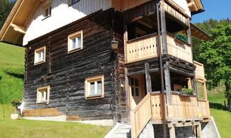 A traditional wooden house with a spacious balcony and several windows. It is situated in a green environment under a blue sky.