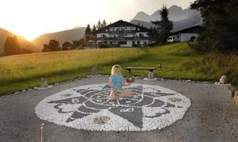 A peaceful environment with a mandala made of stones. A person is meditating in front of a hotel in the background, surrounded by mountains.