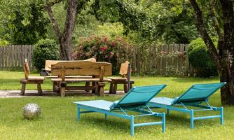 A beautiful garden with two loungers and a wooden table. In the background, green shrubs and a wooden fence can be seen.